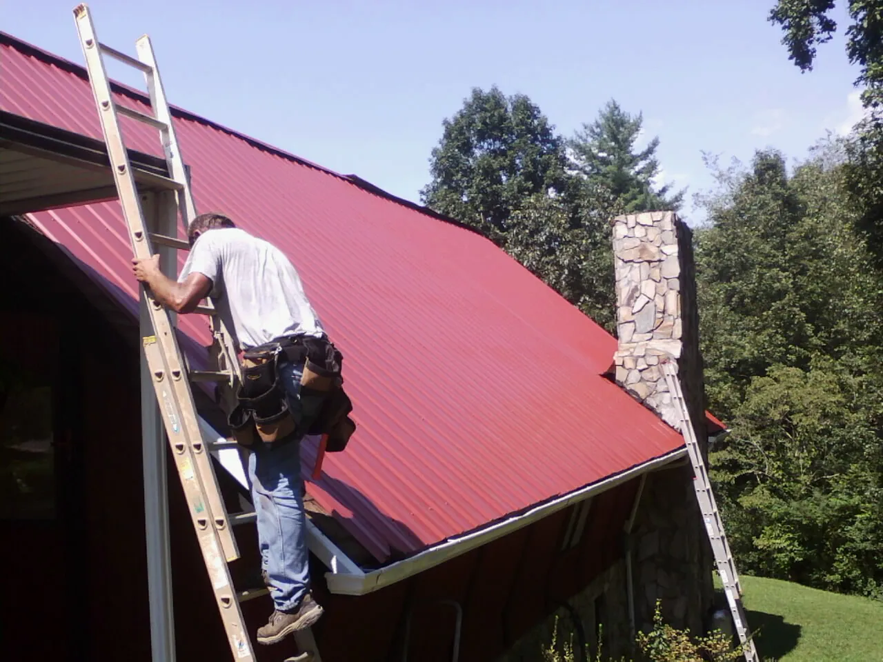 Roofer on a ladder beside a red metal roof and stone chimney — Soffit & Fascia Repair by Roofing Brothers in Strabane